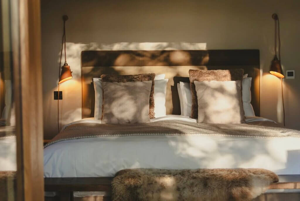 Sunbeams and leafy shadows filter across a beautifully styled king bed with brown and beige pillows, flanked by warm, glowing sconces.