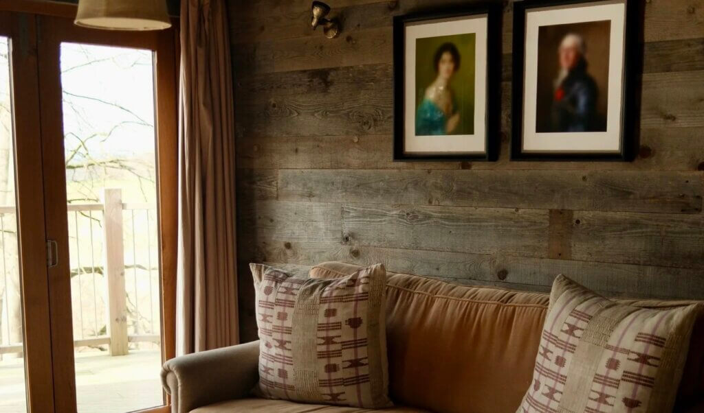 A cosy interior corner featuring a tan sofa with patterned pillows set against a rustic, weathered wood panel wall adorned with two framed portraits.