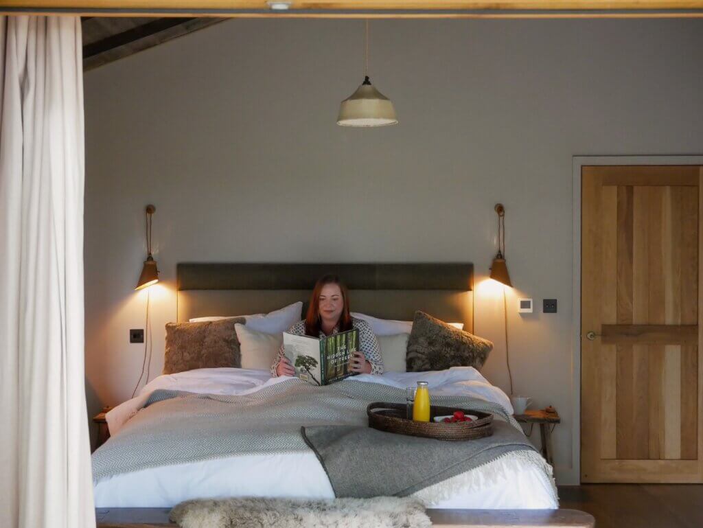 A woman relaxes in a large comfortable bed with neutral-toned linens inside a cosy luxury treehouse, reading a book titled The Hidden Life of Trees with a breakfast tray sitting nearby.