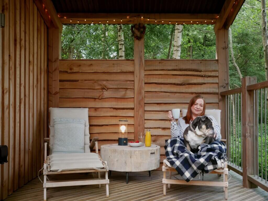 A woman sits on a wooden deck chair wrapped in a plaid blanket, holding a mug and smiling at a bulldog resting on her lap under a covered porch.
