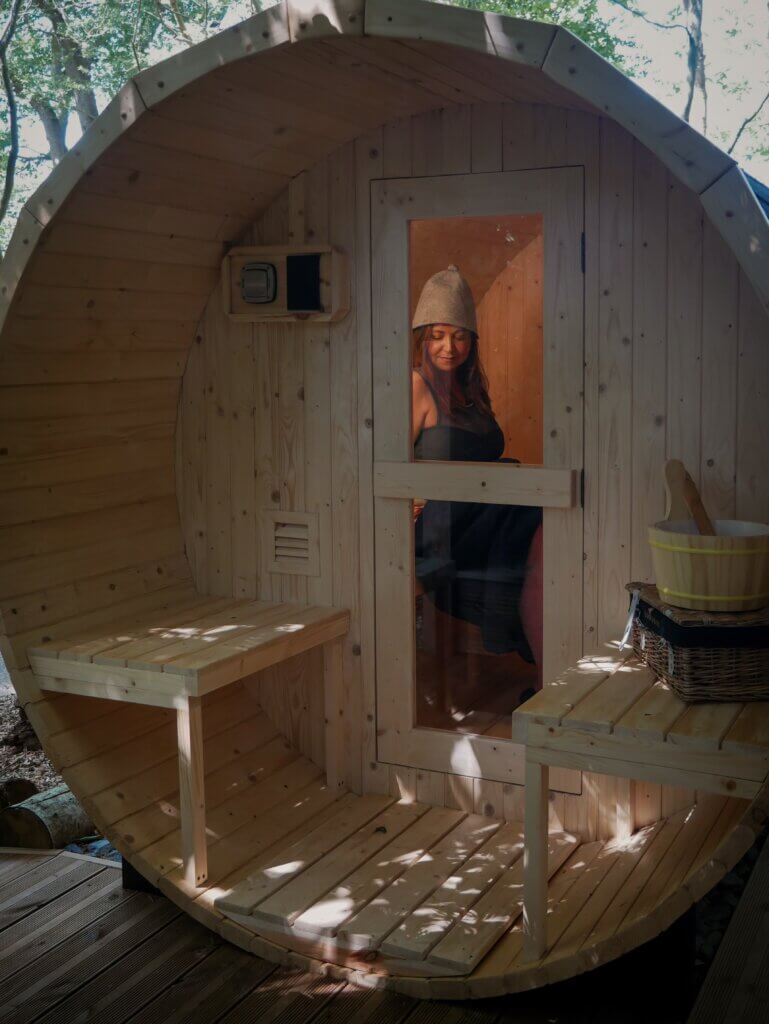A woman wearing a dark swimsuit and a wool sauna hat smiles through the glass door of an outdoor, barrel-shaped wooden sauna.