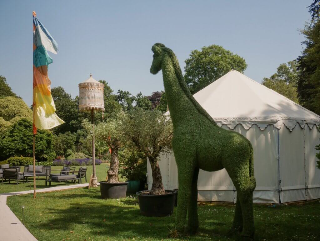 A large grassy lawn featuring a white glamping yurt, a tall green giraffe topiary sculpture, potted olive trees, a colourful flag banner, and a decorative tasseled umbrella pole under a clear blue sky.