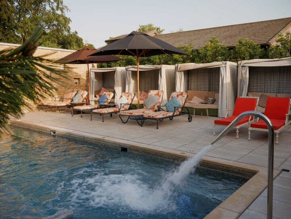 A tranquil resort swimming pool area featuring a water fountain jet spouting into a bubbling pool in the foreground, with patterned lounge chairs and luxury cabanas lined up under dark umbrellas along the tiled patio.