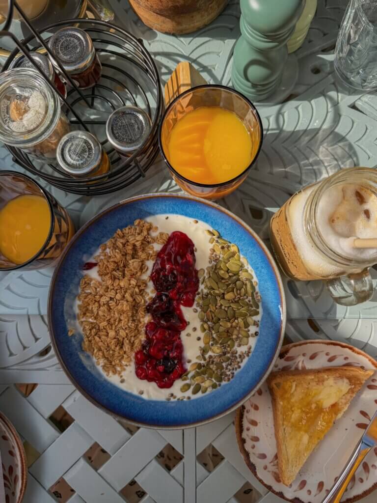 A top-down view of a breakfast table showing a blue ceramic bowl filled with yogurt and divided rows of granola, berry compote, and pumpkin seeds, surrounded by glasses of orange juice, an iced coffee jar and a slice of toast with jam.