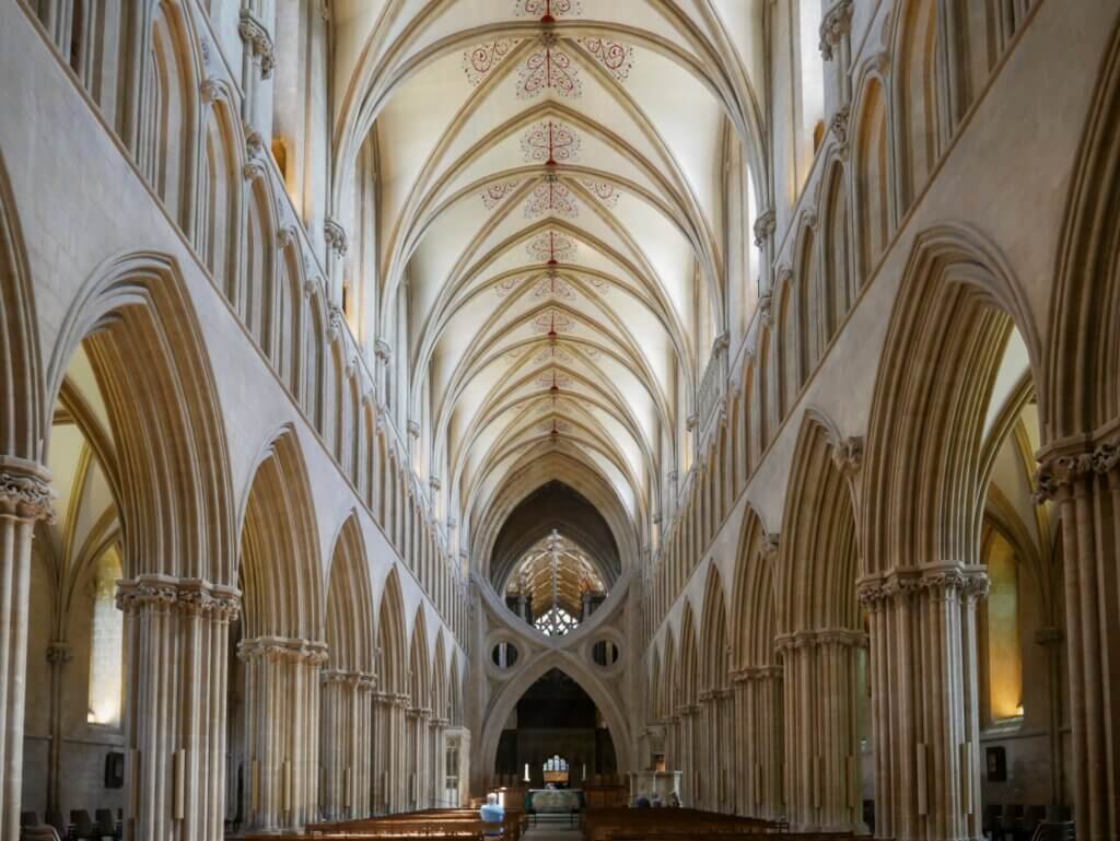 An interior view looking down the long nave of Wells Cathedral, featuring towering stone columns that curve into pointed Gothic arches. High overhead, a vaulted ceiling is decorated with delicate red pattern details along the central ridge. In the distance, the cathedral's famous structural "scissor arch" frames the choir area.