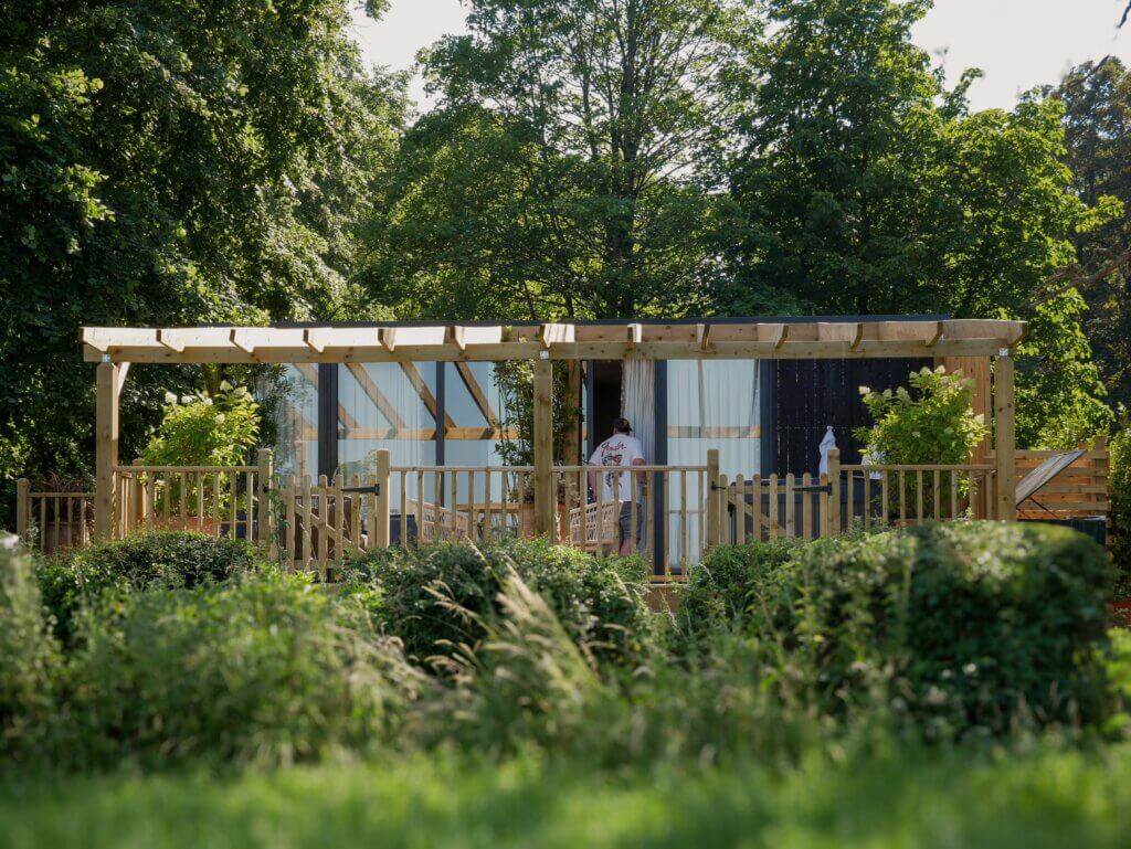 A wide shot of a modern cabin with a wooden pergola and fencing nestled behind lush green bushes and trees, with a person in a white t-shirt visible on the deck.