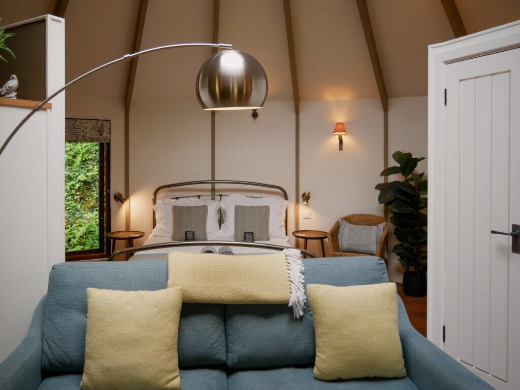 A neatly arranged living room area inside a treehouse, highlighting a blue sofa with yellow throw pillows positioned in front of a metal bed frame with crisp white linens.