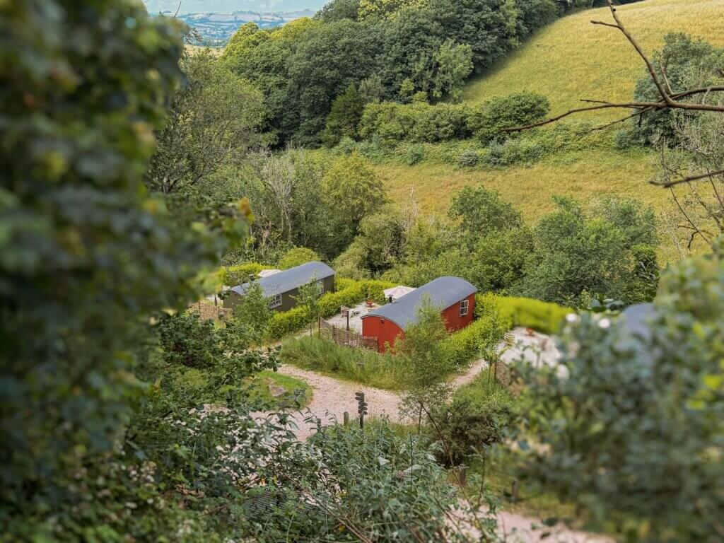An elevated, scenic view looking down onto several rustic red-and-grey glamping cabins tucked away in a verdant rolling green valley under a partly clouded sky.