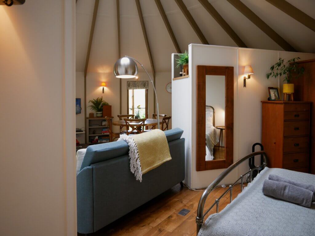 An interior shot of a luxury treehouse showing a cosy blue sofa dividing a living space from a bedroom area featuring a wooden dresser and a large mirror.