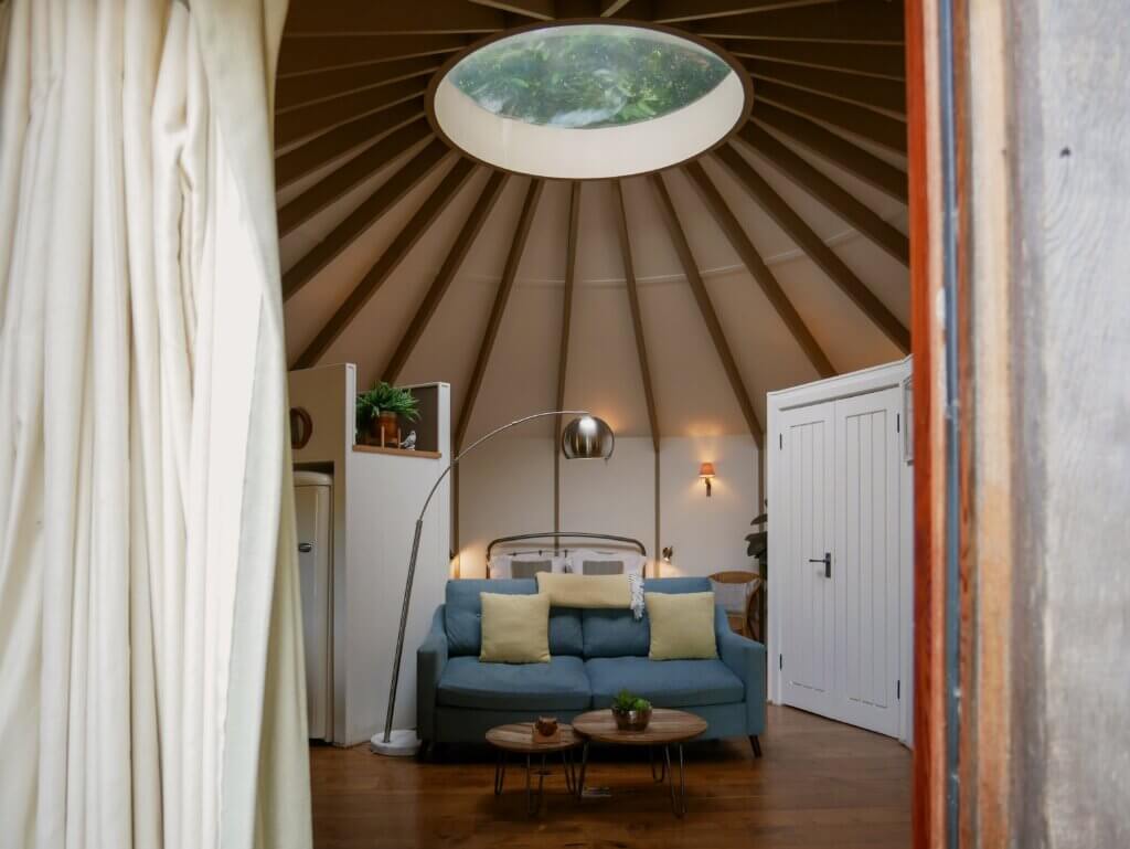 A wide view into the interior of a treehouse, revealing a high, vaulted ceiling with wooden rafters radiating outwards from a central circular skylight that looks out onto green tree leaves.