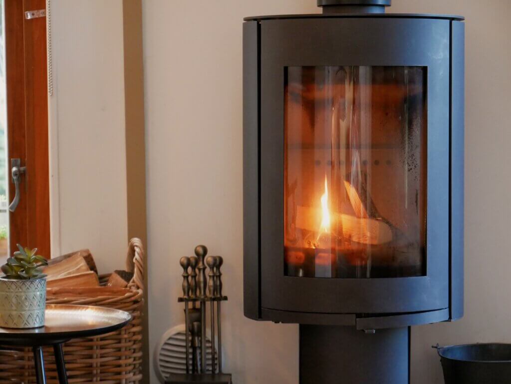 A close-up view of a modern, cylindrical black wood-burning stove with a roaring orange fire visible through its curved glass window.