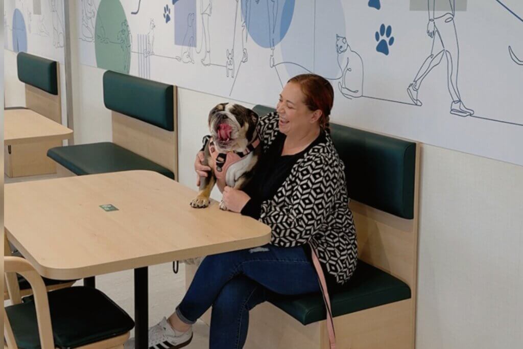 A woman with reddish-brown hair tied back sits on a green-cushioned booth at a wooden table, smiling and hugging an English Bulldog. The bulldog, wearing a pink harness, is sitting on the bench next to her with its mouth wide open in a big yawn. The background features a white wall with blue line art doodles of people walking pets and animal paw prints.