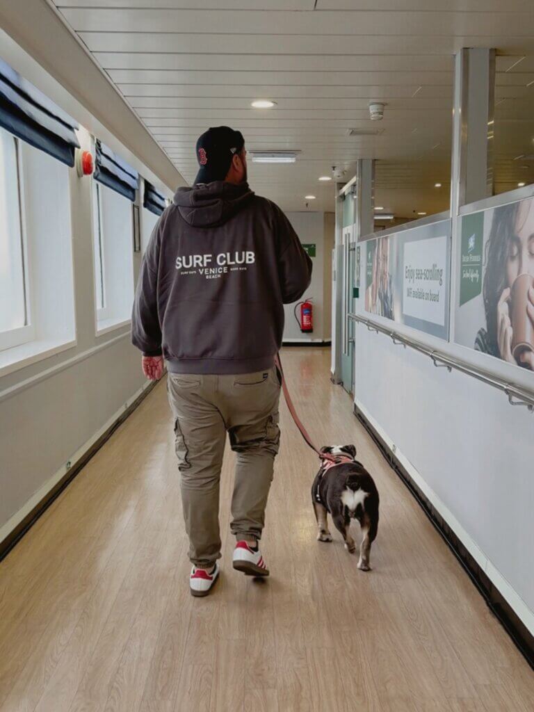 A full-length shot from behind of a man walking a small English Bulldog on a leash down a long corridor of the Irish Ferries ship Innisfree. The man is wearing a dark grey hoodie with "SURF CLUB VENICE BEACH" printed on the back, khaki cargo pants, a black baseball cap, and white sneakers with red stripes. The corridor has light wood-patterned flooring, windows on the left wall with dark blinds, and advertisements on the right wall.