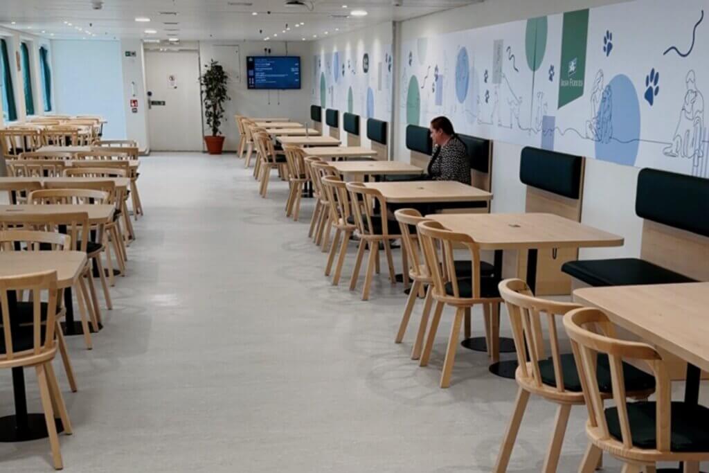 An indoor seating area onboard the Pet Den on Irish Ferries, featuring rows of light-coloured wooden tables and chairs. Along the right wall, there is a booth seating with dark green cushions set against a large mural decorated with paw prints, line drawings of people with pets, and an Irish Ferries logo. A single person sits at one of the booth tables in the distance.