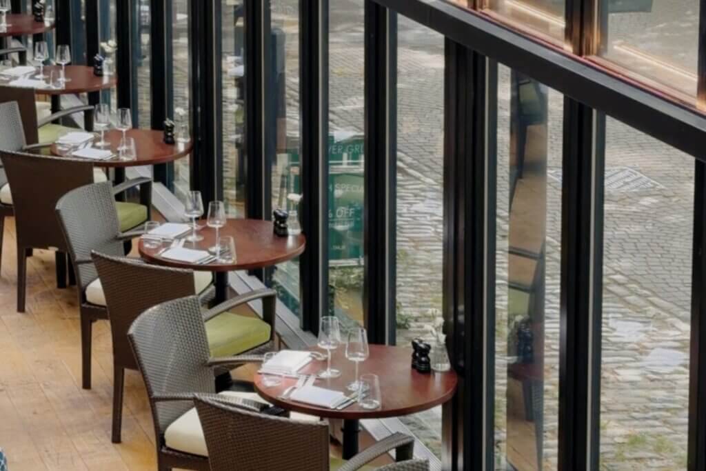 A high-angle view of a restaurant dining area, in The Bristol Hotel, next to floor-to-ceiling glass windows, showing small, round wooden tables set with wine glasses and woven armchairs.