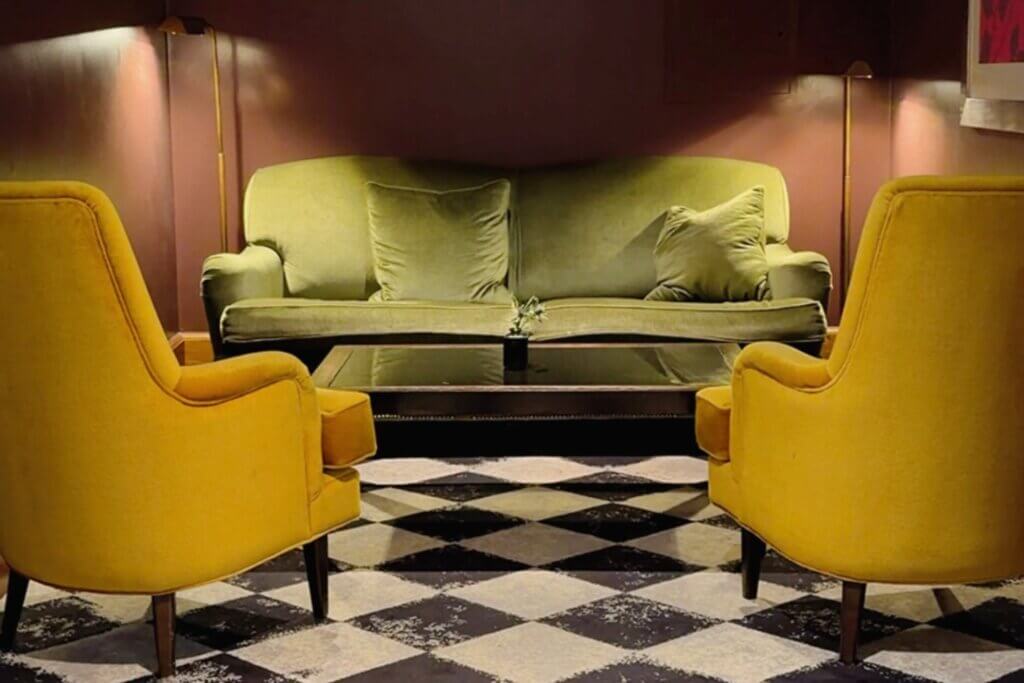 A cosy seating vignette in a dimly lit lounge showing an olive-green velvet sofa flanked by two mustard-yellow armchairs, arranged around a coffee table on a black-and-white checkered rug.