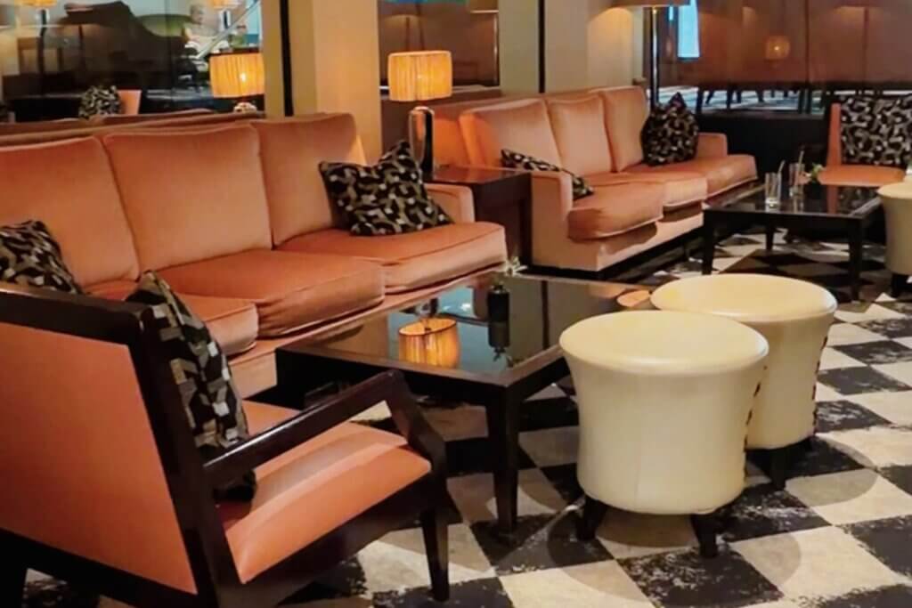 A lounge area in The Bristol Hotel featuring plush peach-coloured sofas, dark wood coffee tables, matching armchairs with patterned pillows, and round cream-coloured ottomans over a black-and-white checkered carpet.