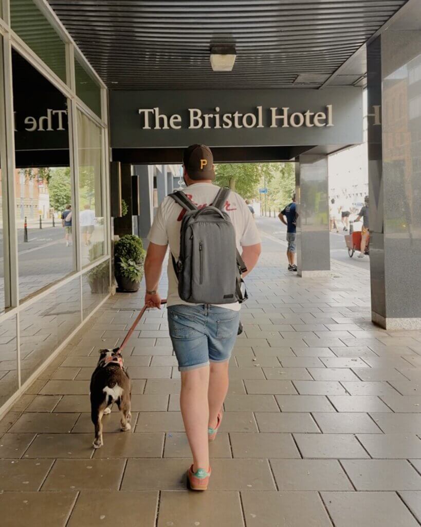 Rear view of a person wearing denim shorts, a white t-shirt, a grey backpack, and a brown baseball cap, walking a bulldog on a leash along a covered tiled walkway outside "The Bristol Hotel."