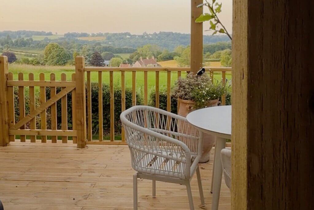 A view from a wooden porch looking out over a small gate and railing toward a vast green countryside with rolling hills and a distant house under a soft, overcast sky.