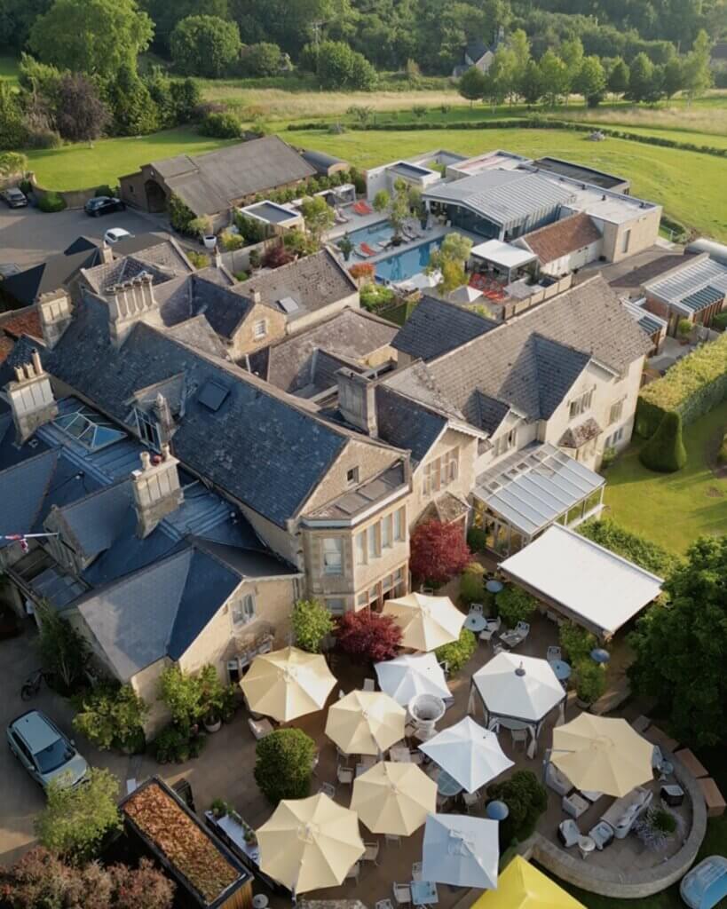 A wide aerial view of a sprawling stone country estate with dark tiled roofs, showing a patio filled with dining tables and cream-coloured umbrellas in the foreground, and the luxury pool area extending into the background.