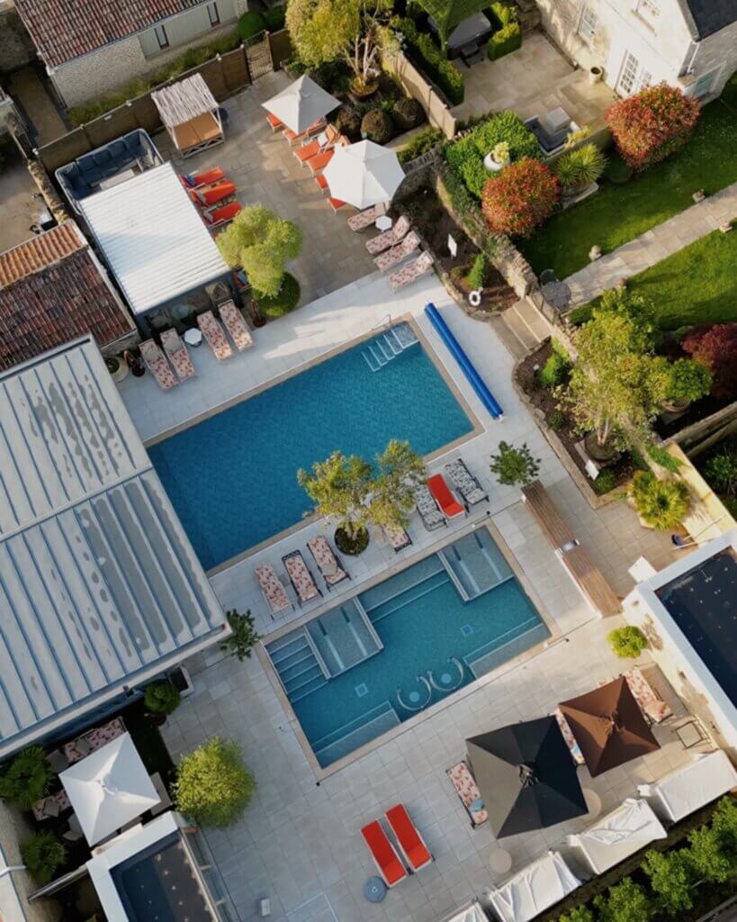 A crisp, top-down aerial view of a luxury hotel patio and pool area, at the Homewood Bath, featuring a long rectangular swimming pool, a smaller segmented hot tub pool, numerous lounge chairs, and scattered patio umbrellas.
