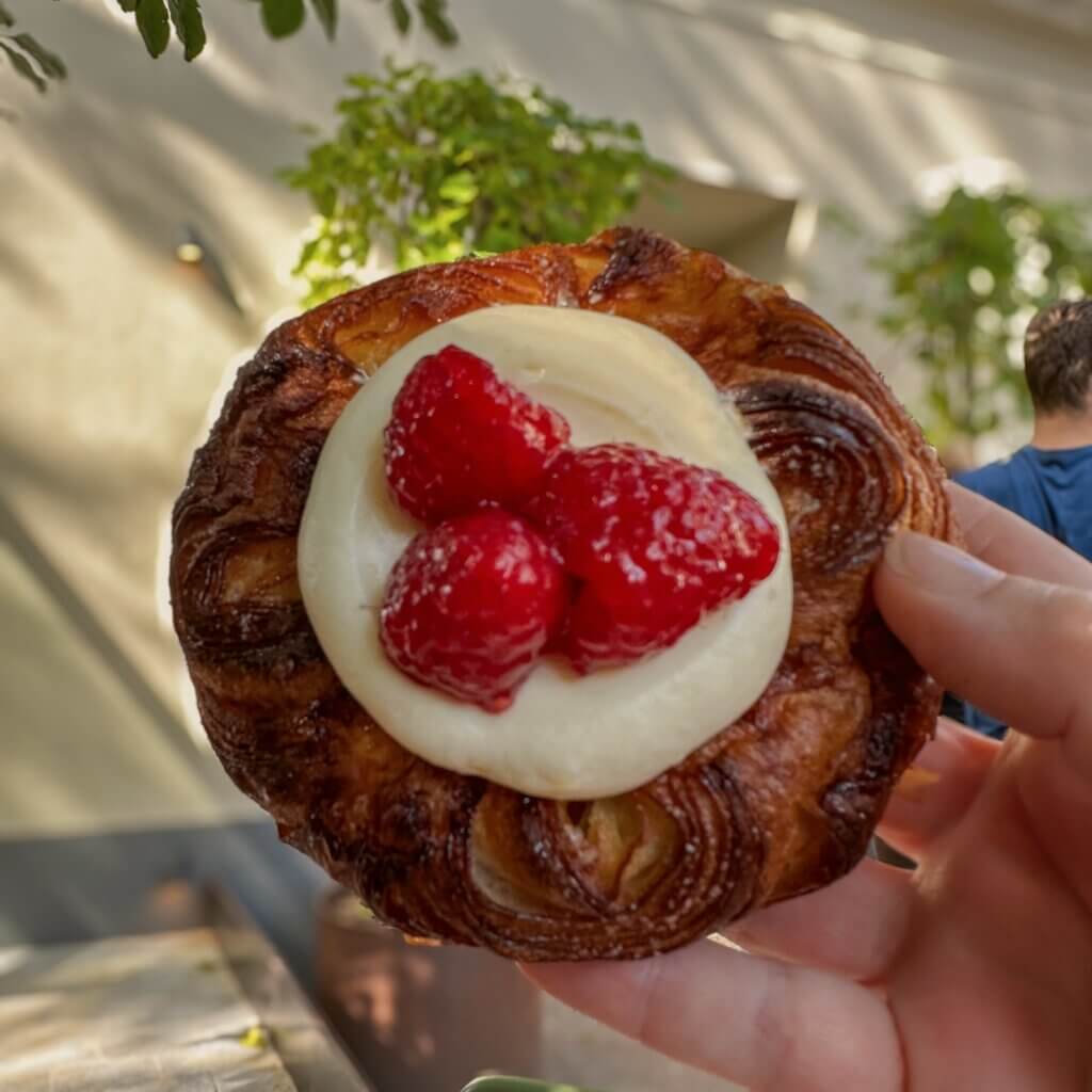 Hand holding a flaky golden-brown Danish pastry topped with vanilla pastry cream and fresh red raspberries outdoors in Copenhagen.
