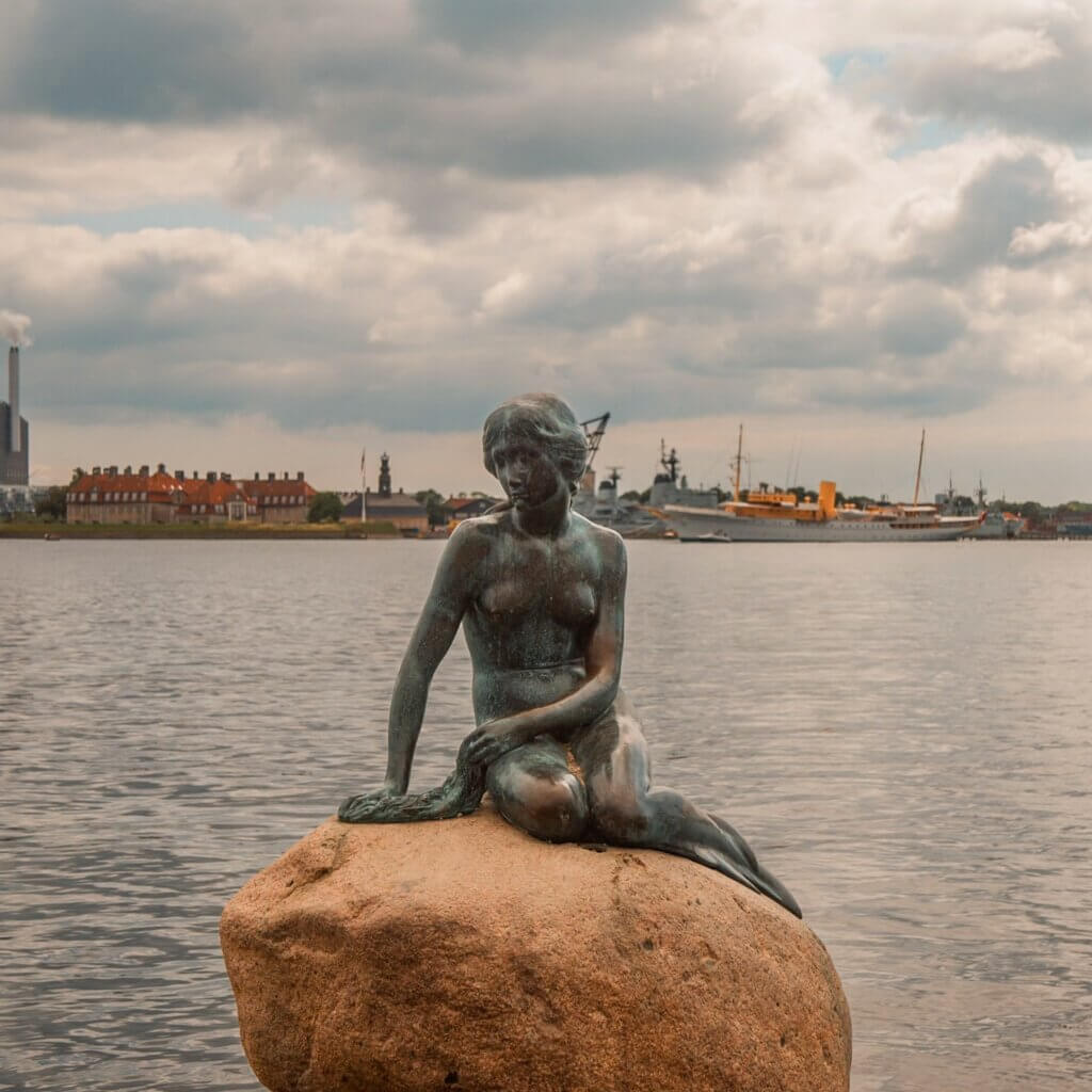 The iconic bronze Little Mermaid statue sitting on a rock in the water at Langelinie promenade, with ships and coastal buildings across the harbour in Copenhagen.