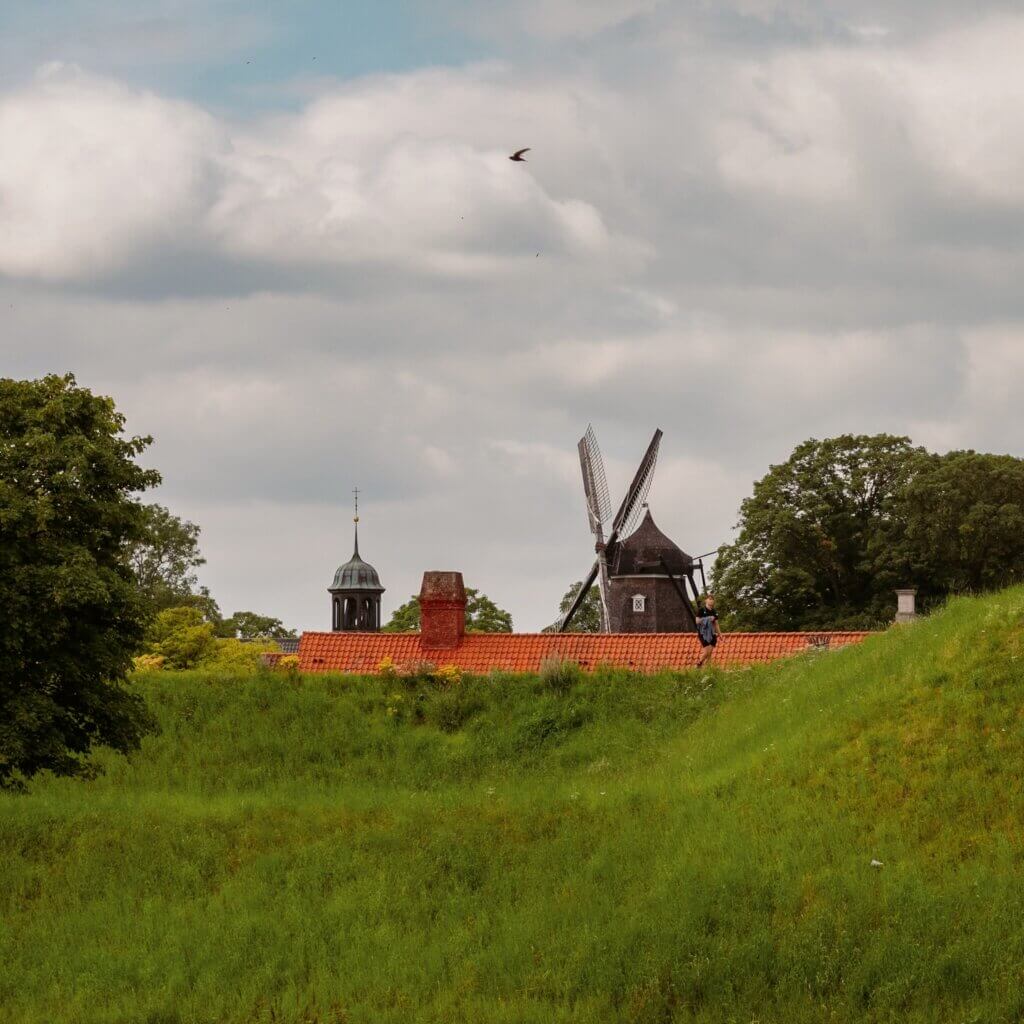 Scenic view across grassy earthwork ramparts toward terracotta rooftops, a spire, and the historic wooden Kastellet Windmill under a cloudy sky in Copenhagen.