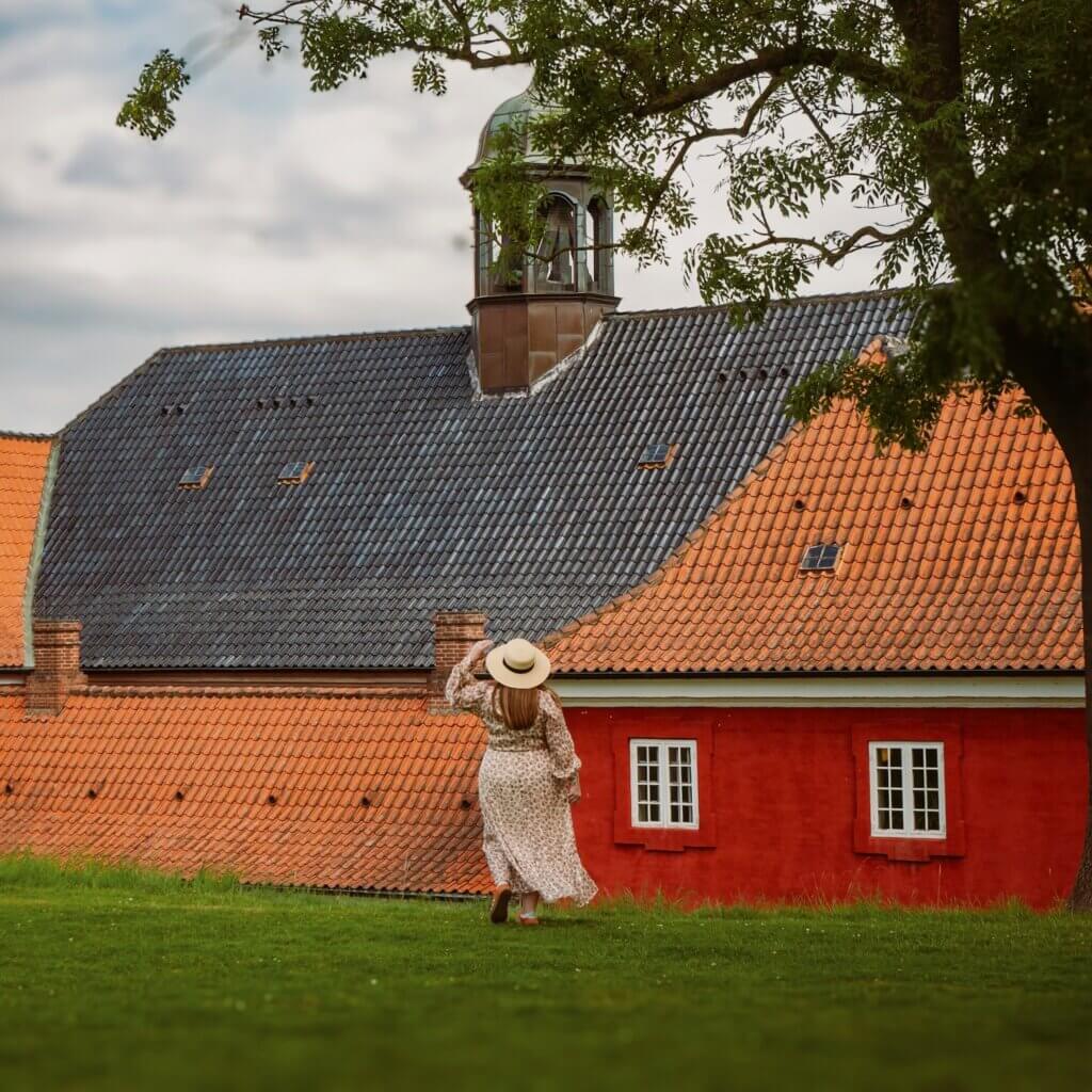 A woman in a long floral dress and straw hat walking along a grassy hill towards a historic red building with terracotta tile roofing and a copper bell tower cupola in Copenhagen.