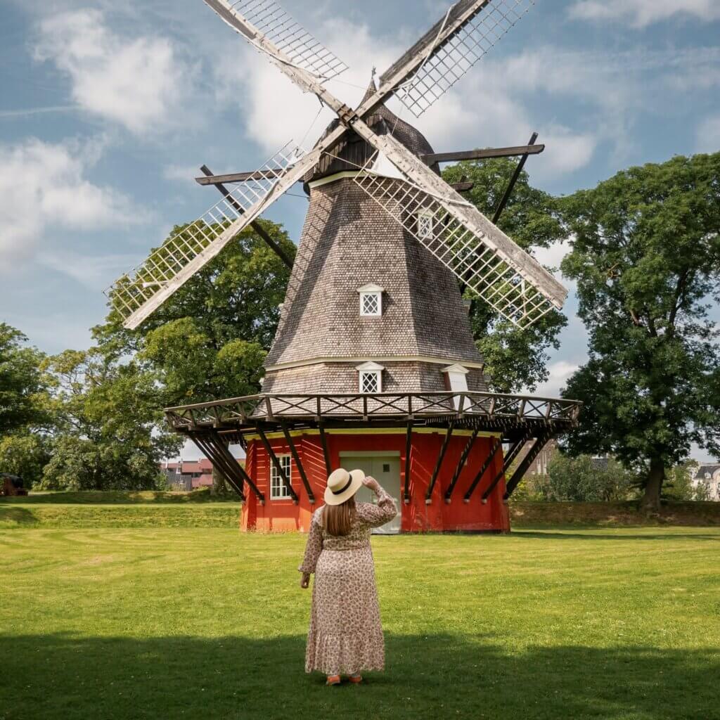 A woman in a floral dress and straw hat standing on a grassy lawn, looking up at the historic red and wooden Kastellet Windmill under a bright blue sky in Copenhagen.