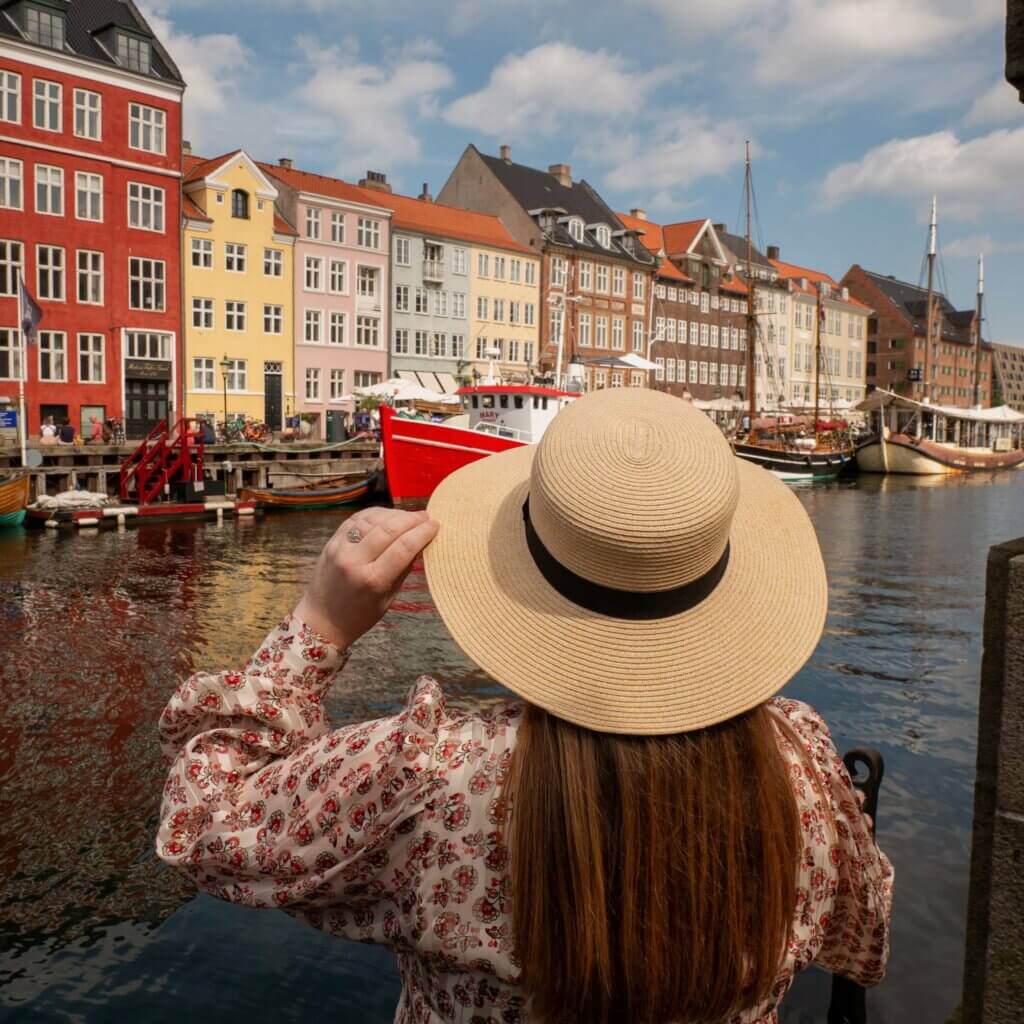 Back view of a woman holding her straw hat while looking across the water at colorful historic harbour houses and boats along Nyhavn in Copenhagen.