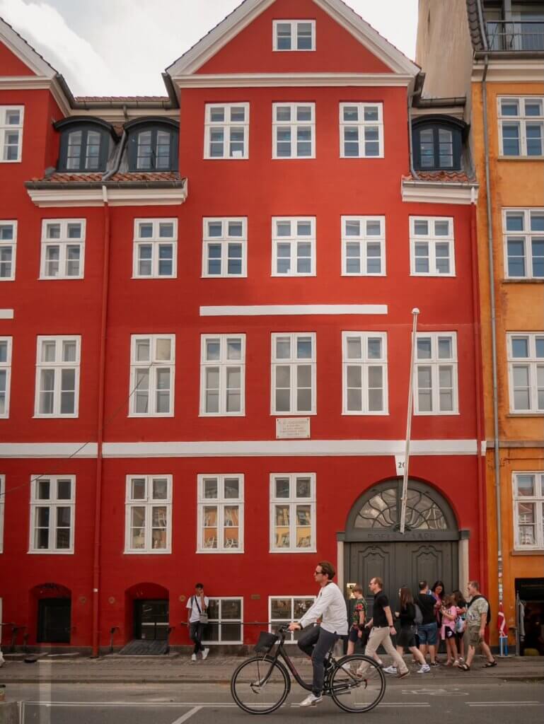A man riding a bicycle past a vibrant red historic building facade with white-framed windows and a grey arched doorway along a street in Copenhagen.