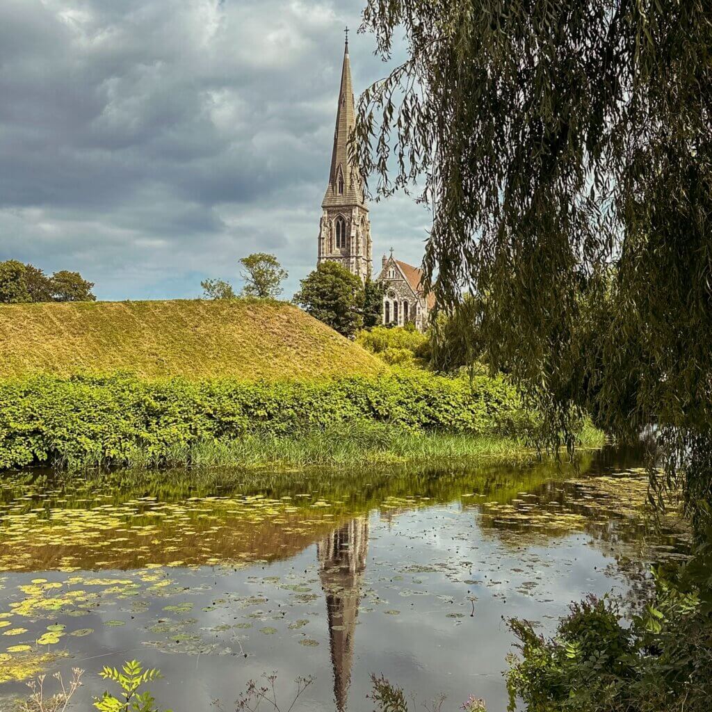 St. Alban's Church steeple reflecting in the calm waters of the Kastellet moat, framed by lush green banks and weeping willow branches in Copenhagen.