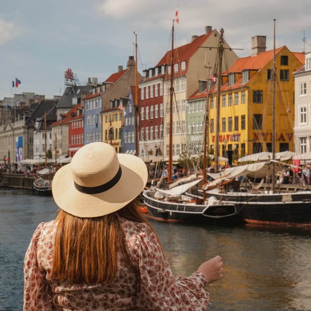 A woman in a straw sunhat and floral blouse enjoying the view of colorful historic buildings and wooden boats along Nyhavn canal in Copenhagen.