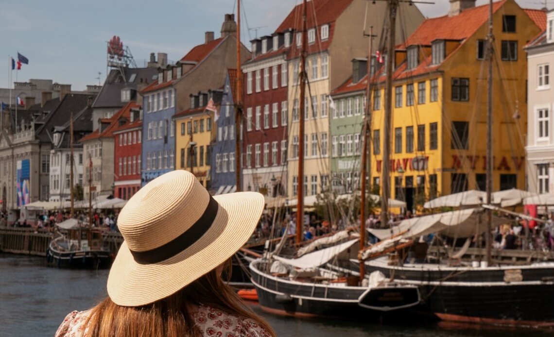 Back view of a woman wearing a straw sunhat and floral dress looking across the water at the iconic colourful townhouses and wooden sailing ships at Nyhavn harbour in Copenhagen.