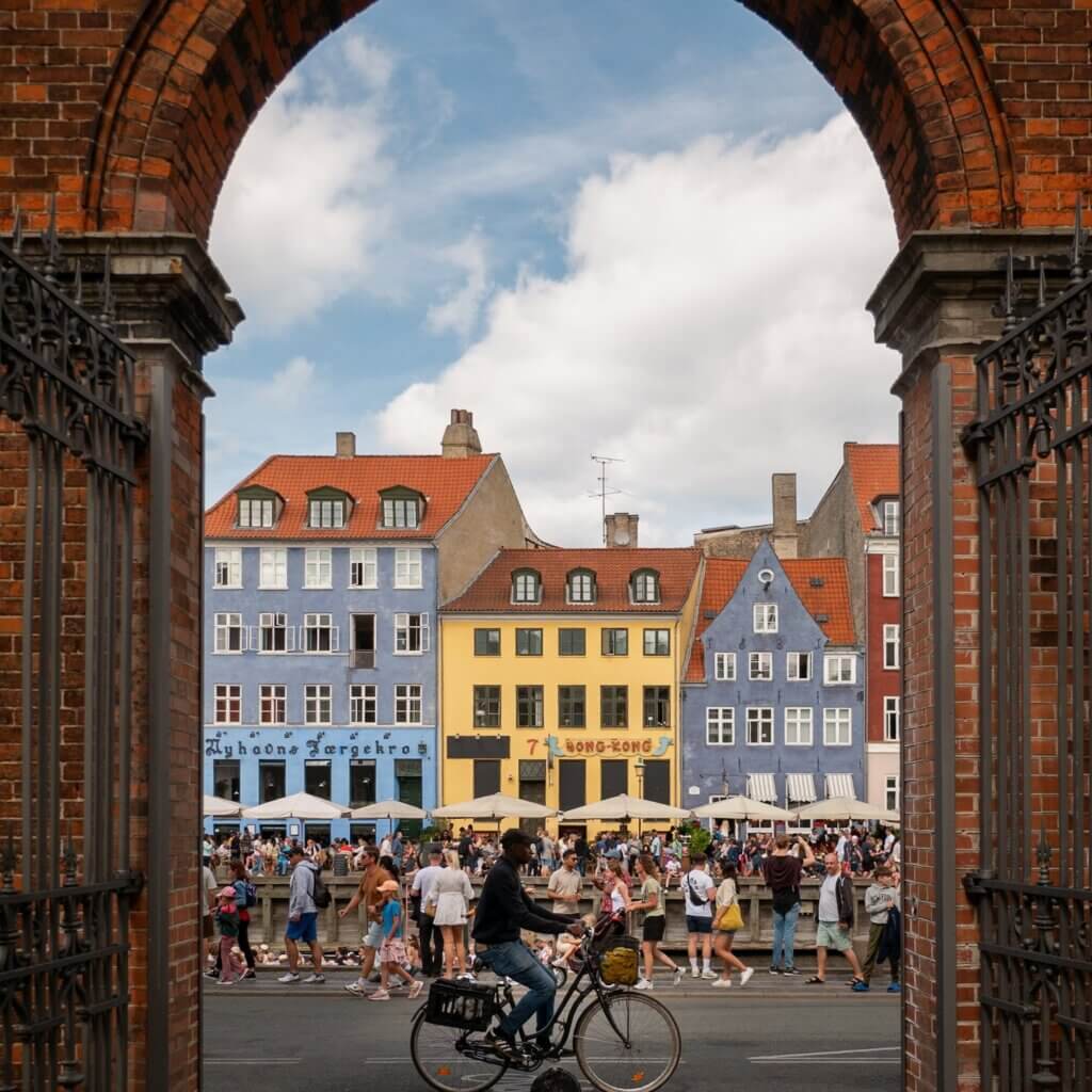 View framed by a red brick archway showing a cyclist riding past busy crowds and colorful historic harbor buildings along Nyhavn in Copenhagen.