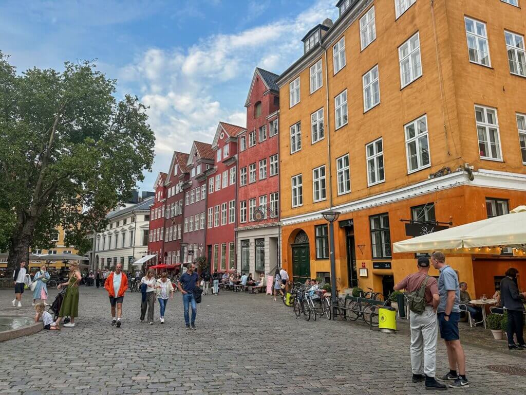 Pedestrians walking across a historic cobbled square surrounded by colorful yellow and red historic buildings, cafe terraces, parked bicycles, and trees in Copenhagen.