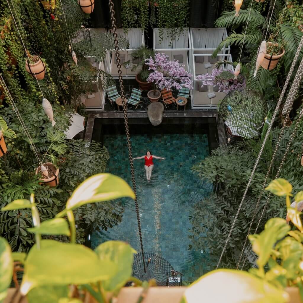 High-angle view of a woman floating in a red swimsuit inside a turquoise pool, surrounded by lush tropical plants, hanging vines, and Balinese-style daybeds at Manon Les Suites hotel in Copenhagen.