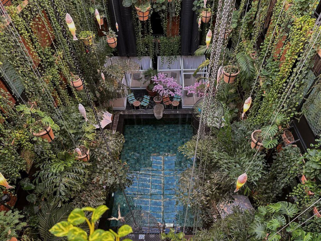 Looking down into a lush indoor atrium pool surrounded by dense green foliage, hanging terracotta planters, floating light fixtures, and Balinese-style daybeds at Manon Les Suites hotel in Copenhagen.