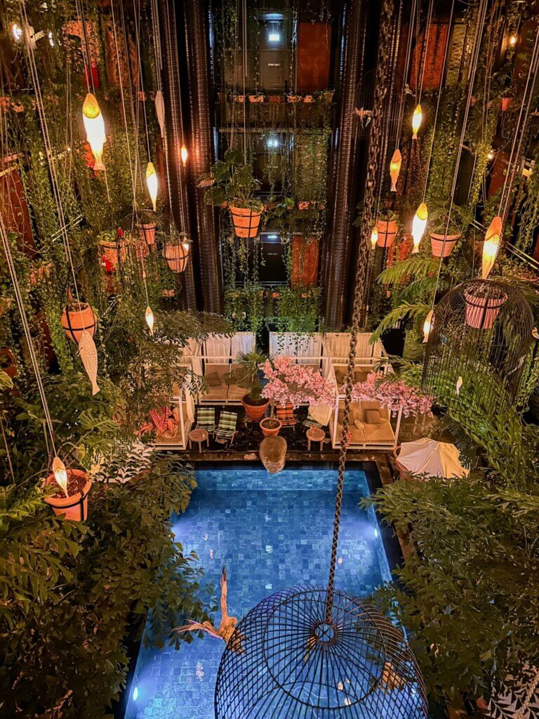 Atmospheric night view looking down into an illuminated indoor pool atrium surrounded by tropical greenery, glowing hanging fish lanterns, and lounge areas at Manon Les Suites hotel in Copenhagen.