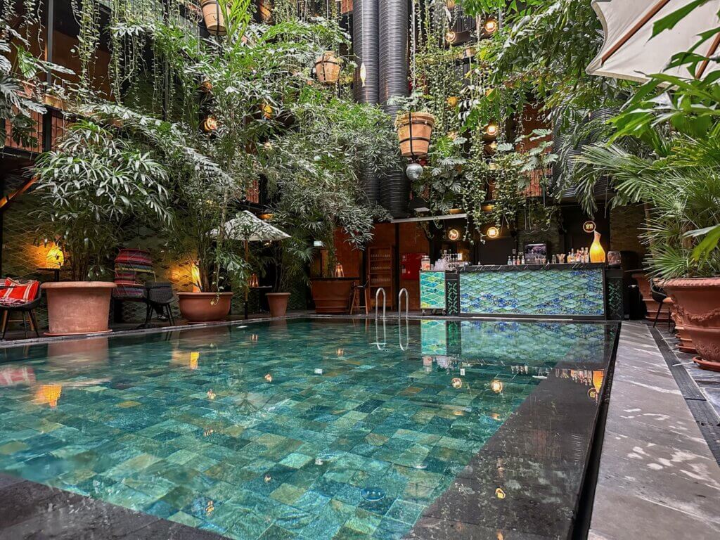 Ground-level view of a clear turquoise indoor pool surrounded by large potted plants, a tiled pool bar, and dangling greenery inside Manon Les Suites hotel in Copenhagen.