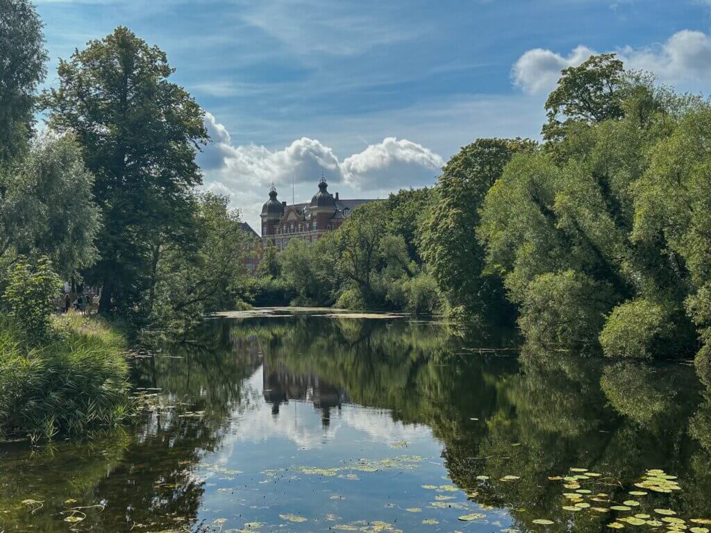 A quiet canal surrounded by lush green trees and lily pads, reflecting a grand historic brick building with domed towers under a blue sky in Copenhagen.