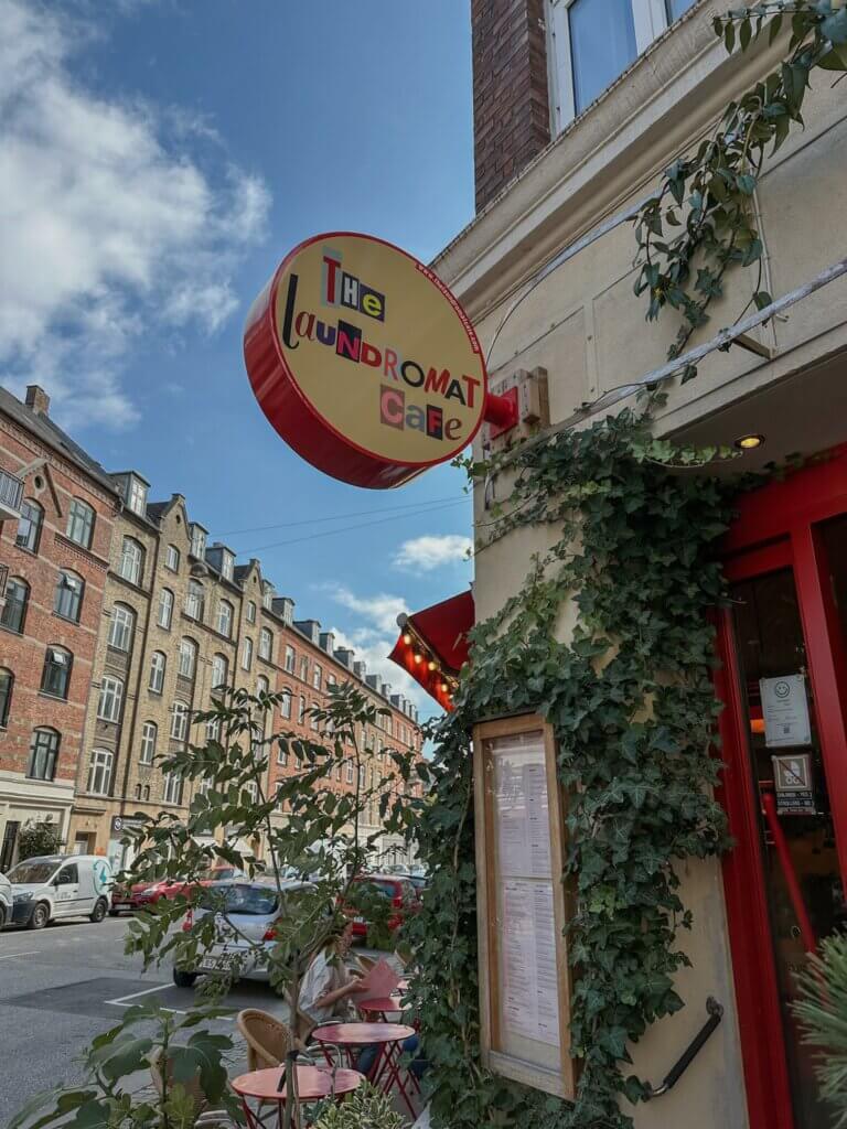 A circular yellow sign with colorful lettering reading "The Laundromat Cafe" hanging on an ivy-covered brick building corner along a cobblestone street in Copenhagen.