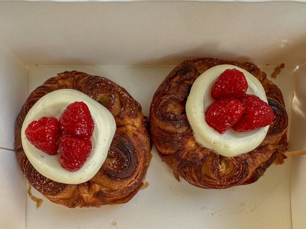 Top-down view of two flaky, golden-brown laminated Danish pastries served in a cardboard takeaway box, each topped with smooth vanilla cream and fresh red raspberries.