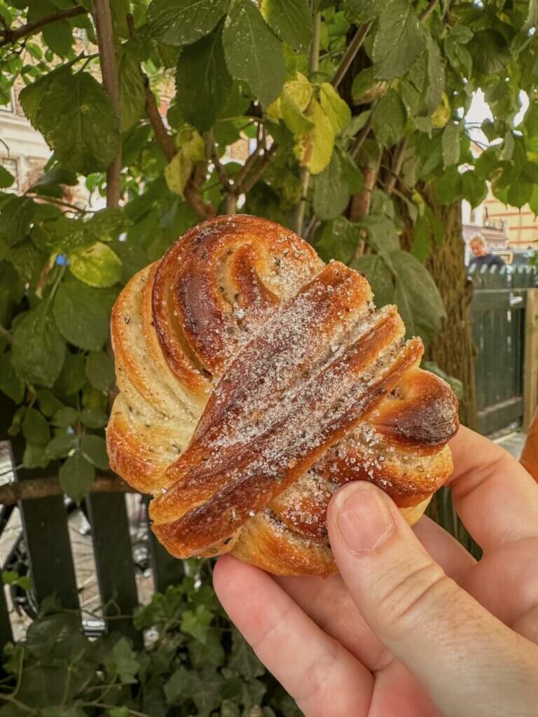 Hand holding a freshly baked, twisted cardamom bun dusted with sugar against a backdrop of green tree foliage in Copenhagen.