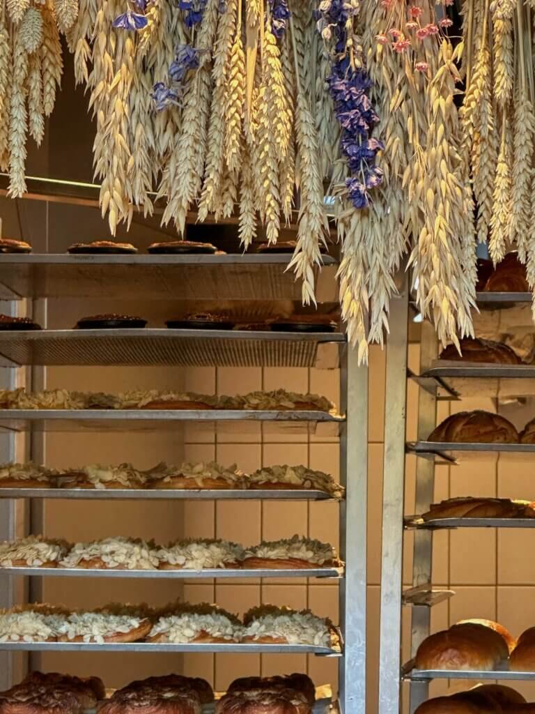 Trays of fresh almond croissants and loaves of bread resting on metal baking racks beneath hanging dried wheat stalks and purple flowers inside a Copenhagen bakery.
