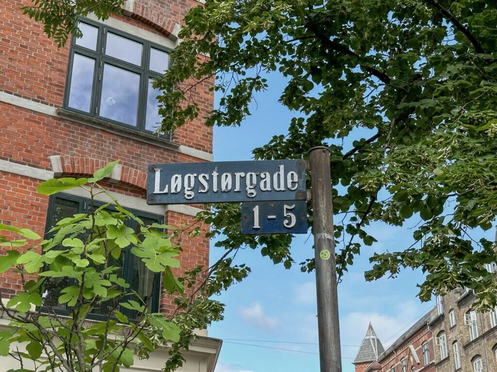 Blue enamel street sign reading "Løgstørgade 1-5" mounted on a pole, framed by leafy green trees and a historic red brick building facade in Copenhagen.