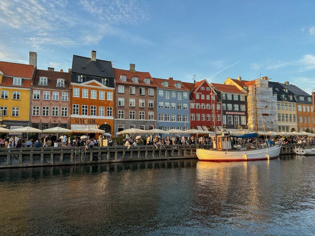 Warm sunlight illuminating a row of colorful historic buildings and outdoor dining patios along the Nyhavn waterfront, with a white boat resting in the canal in Copenhagen.
