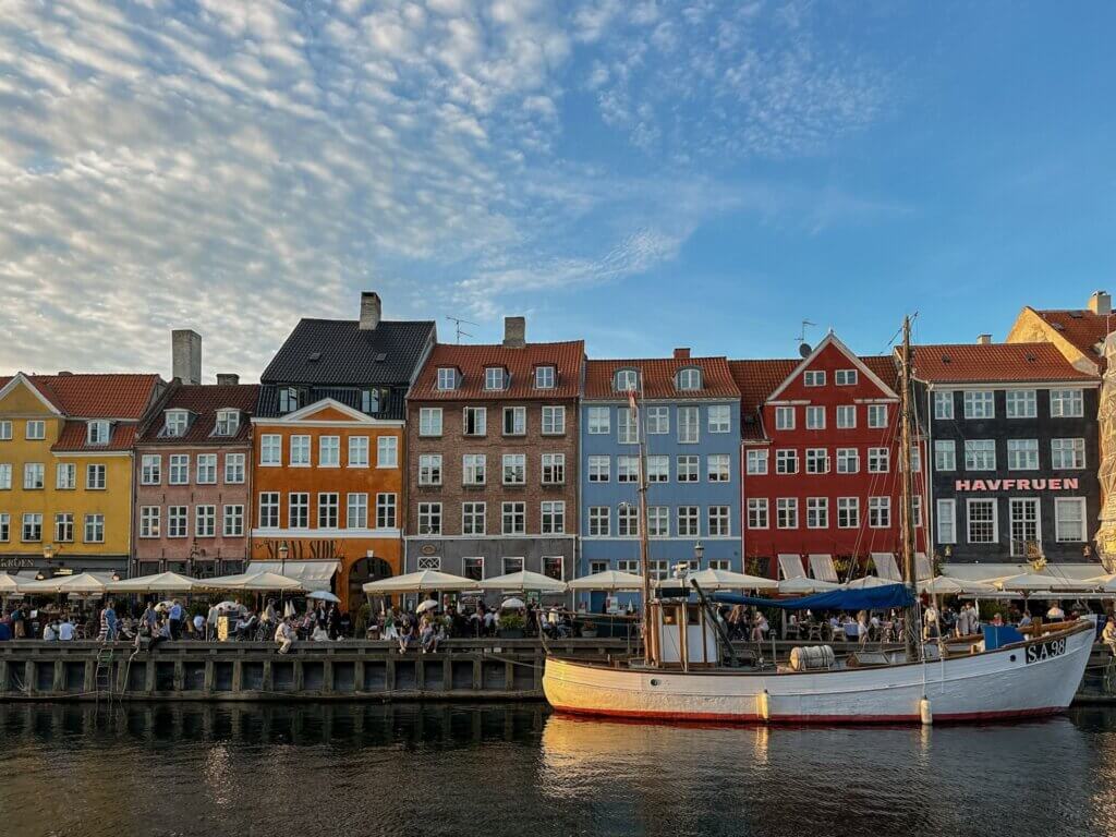 A white fishing boat moored in the canal reflecting in the water, with colorful historic townhouses and busy waterfront cafes under a light-clouded sky at Nyhavn in Copenhagen.