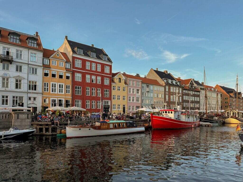 Waterfront view across Nyhavn canal featuring a bright red fishing boat and wooden yacht docked beside a row of colorful pastel and brick buildings under a clear blue sky in Copenhagen.